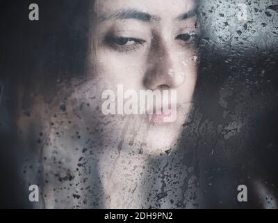 Pensive young Nepalese woman behind wet glass Stock Photo