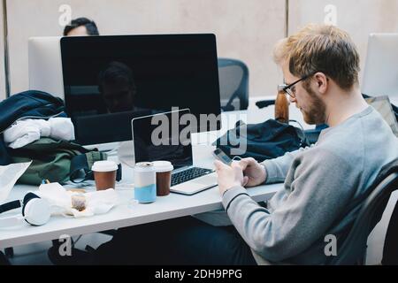 Side view of computer programmer using smart phone at desk in office Stock Photo