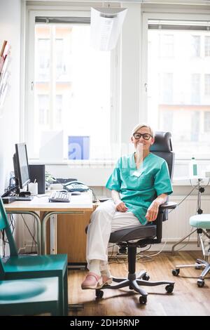 Full length portrait of smiling mature female doctor sitting in clinic Stock Photo