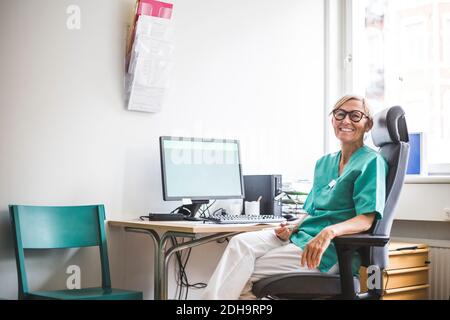 Portrait of smiling mature female doctor sitting on chair at clinic desk Stock Photo