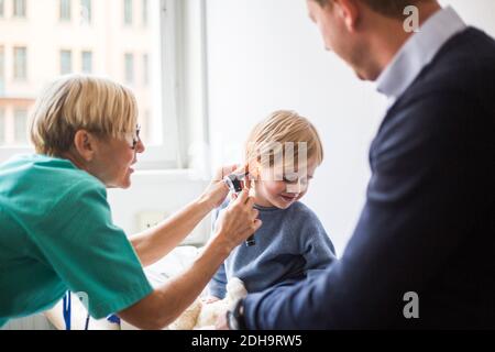 Female doctor examining boy's ear with otoscope while sitting by father in hospital Stock Photo