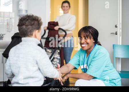 Smiling female nurse shaking hands with boy while mother standing in background at medical clinic Stock Photo