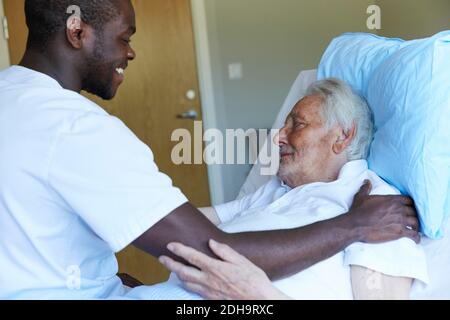 Happy male nurse consoling senior man reclining on bed in hospital ward Stock Photo