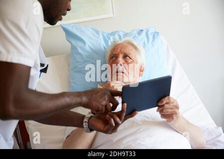 Senior man looking at male nurse while using digital tablet on bed in hospital ward Stock Photo