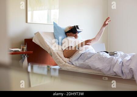 Senior man gesturing while using VR glasses on bed in hospital ward Stock Photo