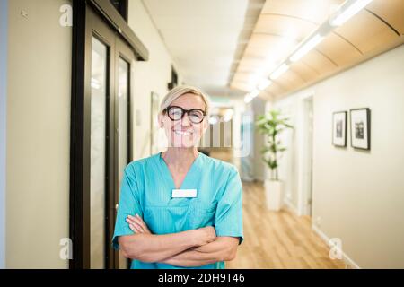 Portrait of mature female healthcare worker with arms crossed standing in hospital corridor Stock Photo