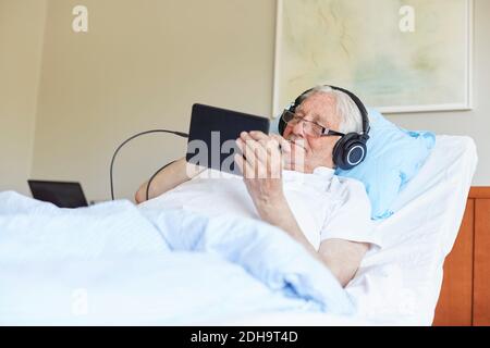 Senior man wearing headphones while using digital tablet on bed in hospital ward Stock Photo
