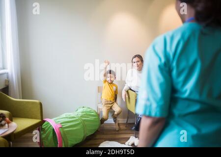 Rear view of doctor while boy with hand raised sitting in hospital Stock Photo