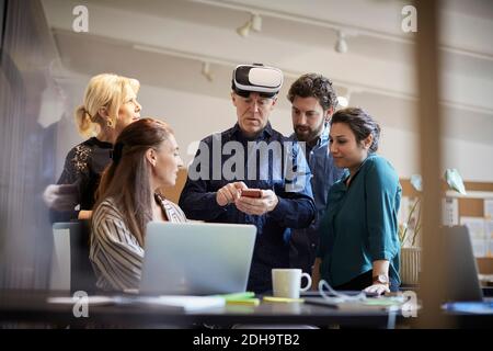 Mature businessman with virtual reality simulator using smart phone while standing amidst colleagues Stock Photo