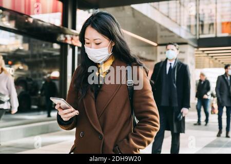 Woman with face mask walking in street using smartphone Stock Photo