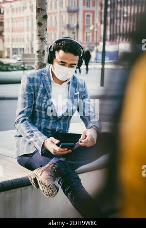 Businessman with smartphone sitting outside with face mask Stock Photo