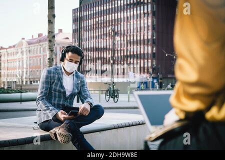 Businessman wearing face mask sitting outside using smartphone Stock Photo