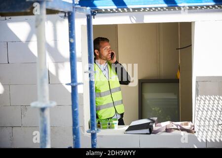Male architect talking on mobile phone while standing at incomplete building Stock Photo