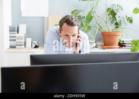 Smiling businessman talking on mobile phone while using computer at creative office Stock Photo