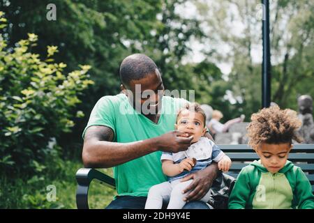 Smiling father feeding baby boy while sitting with son on bench at park Stock Photo