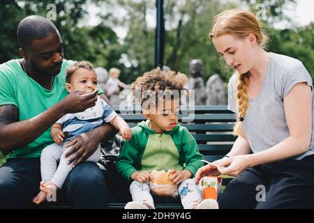 Parents having food with children while sitting on bench at park Stock Photo