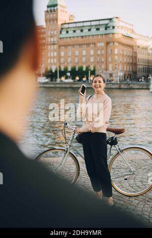 Smiling female with bicycle looking at friend while standing by river in city Stock Photo