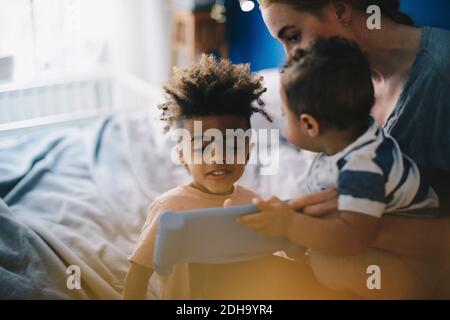 Smiling boy looking at mother with toddler teaching digital tablet at home Stock Photo