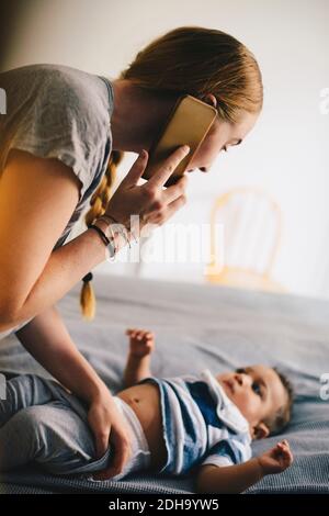 Mother dressing toddler while talking on mobile phone at home Stock Photo