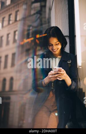 Smiling young woman texting through phone while standing by glass window Stock Photo