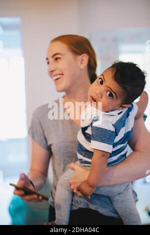 Smiling mother holding mobile phone while carrying son at home Stock Photo