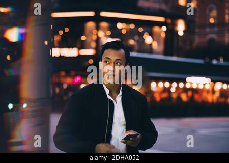 Young man with phone looking away while standing in illuminated city at night Stock Photo