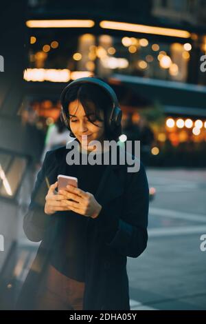 Young woman using mobile phone while wearing headphones in city at night Stock Photo