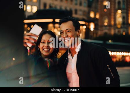 Smiling man and woman taking selfie through phone while standing in city at night Stock Photo