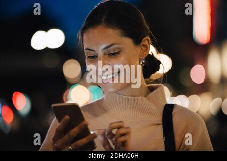 Smiling young woman texting through smart phone in city at night Stock Photo