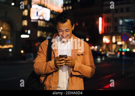 Smiling young man texting through phone while standing in illuminated city at night Stock Photo