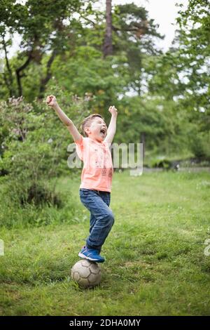 Excited boy shouting with arms raised while standing on soccer ball at back yard Stock Photo