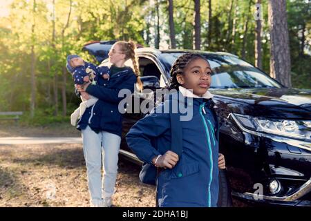 Girl standing against mother carrying baby boy by car amidst trees Stock Photo