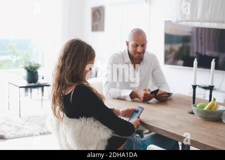 Father and daughter using mobile phone while sitting on table at home Stock Photo
