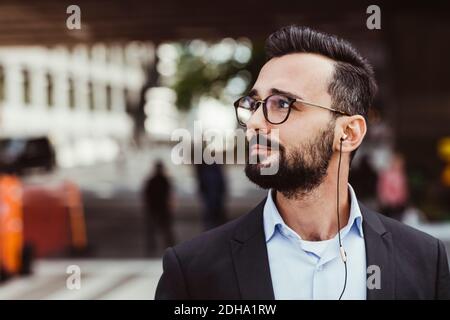 Confident businessman with in-ear headphones looking away while standing outdoors Stock Photo