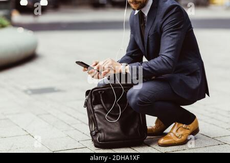 Low section of male entrepreneur using smart phone through in-ear headphones while crouching on street Stock Photo