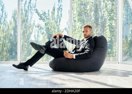 Business man relaxing on bean bag chair with cup of coffee on panoramic windows background in modern office Stock Photo