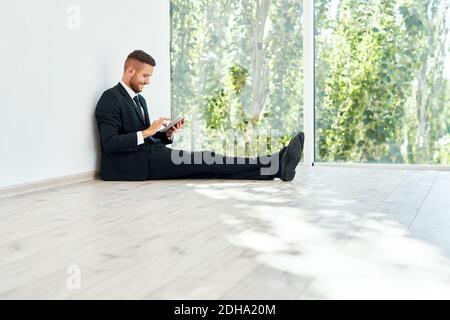 Young smiling businessman sitting on floor in his office and looking at smartphone Stock Photo