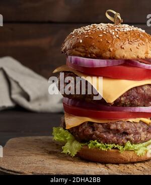 Double cheeseburger with tomatoes, onions, barbecue cutlet and sesame bun on an old wooden cutting b Stock Photo