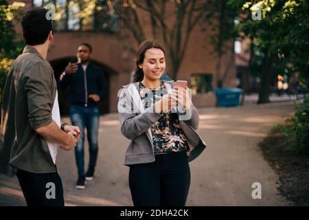 Smiling woman using smart phone while walking with male friends on footpath Stock Photo
