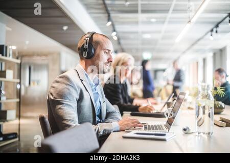Businessman listening headphones while using laptop by colleague at table in office Stock Photo
