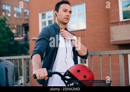 Mid adult man adjusting in-ear headphones while walking with bicycle against building in city Stock Photo