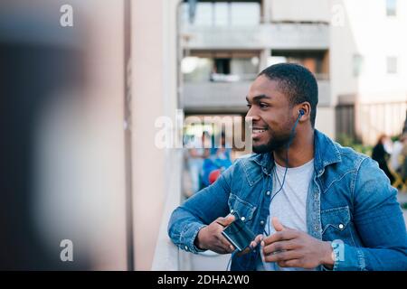 Smiling young man listening to headphones while standing on footbridge in city Stock Photo