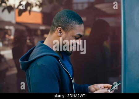 Side view of smiling young man using smart phone against wall Stock Photo