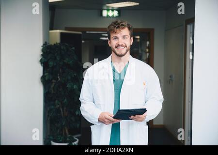 Portrait of confident smiling male doctor standing with digital tablet at hospital corridor Stock Photo