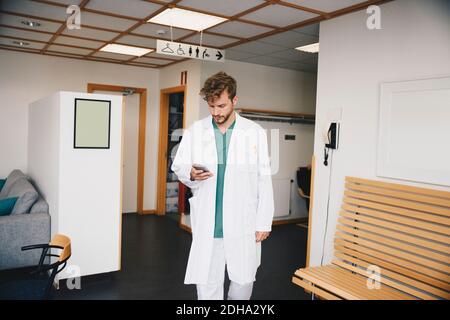Young doctor using smart phone while walking in corridor at hospital Stock Photo