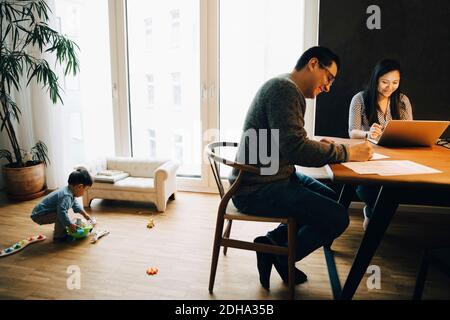 Mother and father working at dining table while son playing with toys in living room Stock Photo