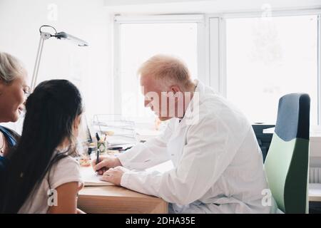 Girl and mother sitting by doctor writing on notepad in office at hospital Stock Photo