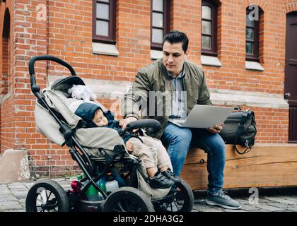 Father using laptop while son sitting in stroller against building in city Stock Photo