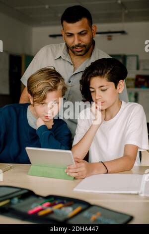 Male tutor with students using digital tablet while sitting in classroom Stock Photo