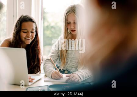 Female student writing in book while sitting by friend at table Stock Photo